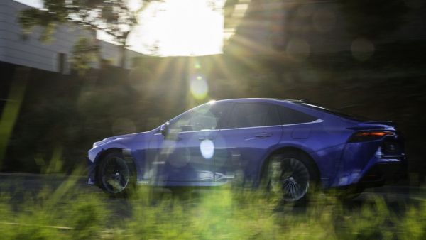 blauer Toyota fährt auf einer Landstraße, neben dem Fahrzeug sind Bäume und Felder. Die Sonne scheint über dem Fahrzeug in die Kamera