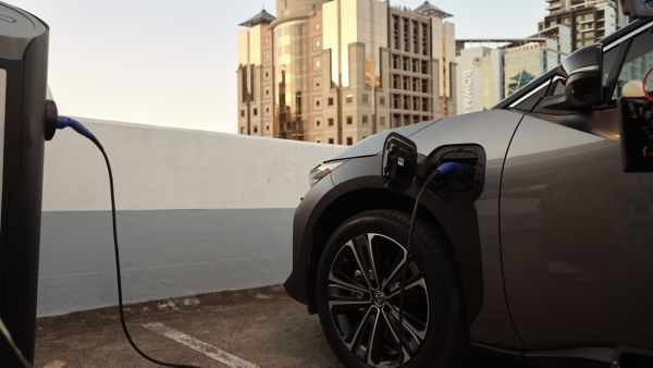 Ein graues Elektroauto wird auf einem Parkdeck bei Sonnenuntergang an einer öffentlichen Ladesäule mit einem blauen Ladekabel geladen. Der Ladeanschluss befindet sich an der Fahrzeugfront, im Hintergrund sind moderne Bürogebäude zu sehen. Die Szene zeigt den Ladevorgang eines E-Autos in der Stadt – eine EV-Ladekarte wird für den Zugang zur Ladestation benötigt.