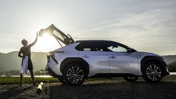 Ein sportlich gekleideter Mann steht bei Sonnenaufgang neben einem modernen weißen Elektro-SUV mit geöffnetem Kofferraumdeckel. Das Fahrzeug ist auf einem Naturparkplatz am See oder Flussufer abgestellt, im Hintergrund sind Berge und Wasser zu sehen. Die Szene vermittelt aktive Elektromobilität in der Freizeit – unterwegs ermöglicht eine Ladekarte für Elektroautos den einfachen Zugang zu öffentlichen Ladesäulen.