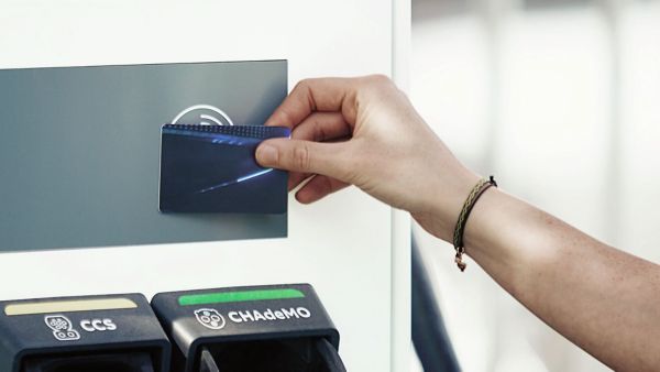 Eine Hand hält eine Ladekarte an ein Gerät, um zu zahlen. Unten sieht man zwei Knöpfe mit der Aufschrift "CCS" und "CHAdeMO"