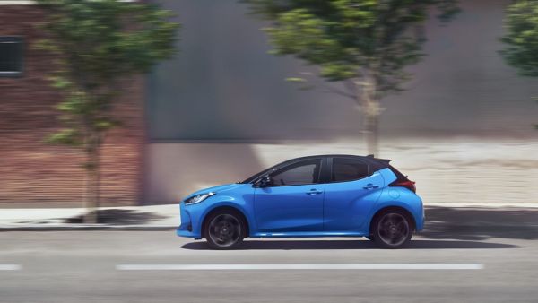 Seitenansicht auf einen blauen Toyota Yaris, der auf einer Straße beschleunigt mit unscharfen Bäumen und Hausfassade im Hintergrund.
