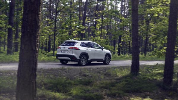 Zu sehen ist ein weißer Toyota SUV mit Hybridtechnik, der durch einen von Bäumen gesäumten Weg inmitten einer ruhigen Waldkulisse fährt