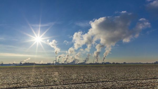 Ein brachliegendes Feld unter blauem Himmel mit Fabriken im Hintergrund, die Rauchschwaden in die Luft abgeben.