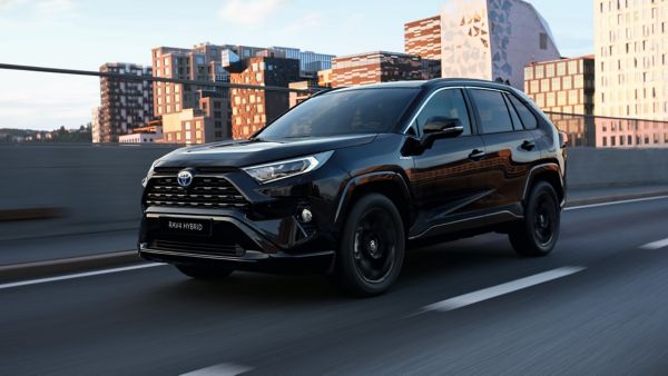 Ein schwarzer RAV4 fährt auf einer Straße mit städtischer Kulisse im Hintergrund.