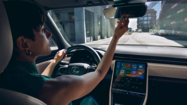 Person sitzt am Steuer eines modernen Autos und greift zum Rückspiegel, während das Fahrzeug auf einer sonnigen Straße zwischen Gebäuden fährt. Auf dem zentralen Display ist eine bunte Benutzeroberfläche mit Apps sichtbar.