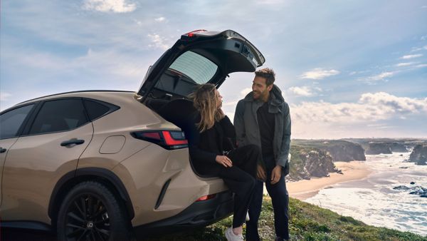 Ein Paar genießt die Aussicht von seinem beigen Lexus NX mit offenem Kofferraum auf einen Strand und Klippen unter blauem Himmel.