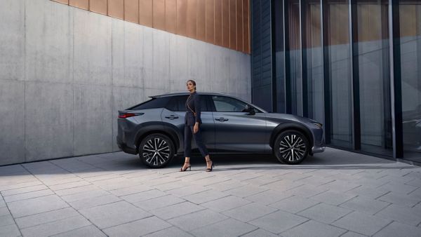 Eine elegant gekleidete Frau steht neben einem grauen Lexus NX vor einer modernen Gebäudefassade aus Beton, Glas und Kupfer.