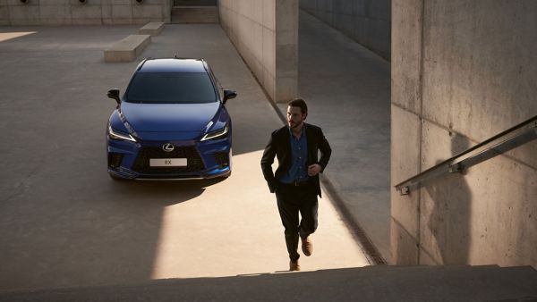 Ein Mann im Anzug läuft eine Treppe hinauf zu einem modernen, blauen Lexus, der in einem minimalistischen, gut beleuchteten Außenbereich geparkt ist.
