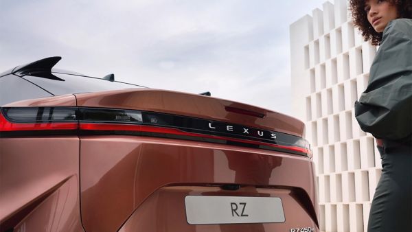 Eine elegante Frau steht neben einem bronzenen Lexus RZ, der sein elegantes Heckdesign und sein unverwechselbares Branding vor einem modernen Hintergrund zur Schau stellt.