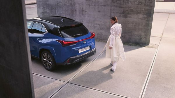 Eine Frau in einem langen weißen Mantel geht von einem blauen Lexus UX 300h weg, der in der Nähe einer modernen Betonstruktur geparkt ist