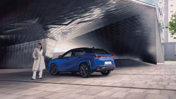 Eine elegante Frau in einem weißen Mantel steht neben einem blauen Lexus UX 300h, der vor einer modernen architektonischen Kulisse geparkt ist.