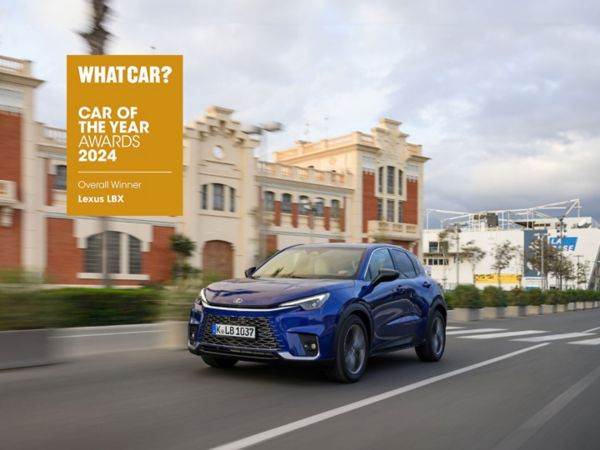 Ein blauer Lexus LBX fährt entlang einer malerischen Straße mit historischen Gebäuden und wurde von What Car? als Auto des Jahres 2024 ausgezeichnet.