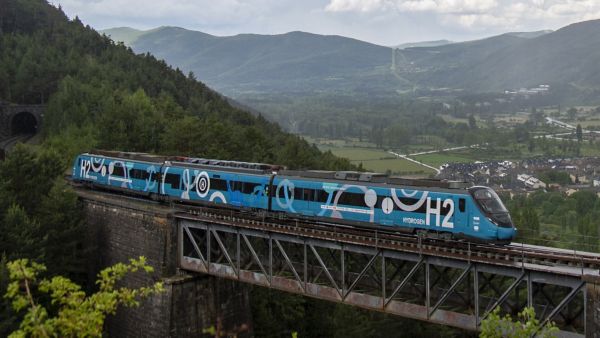 Blauer Zug mit weißem Label "H2 Hydrogen" fährt über eine Stahlbrücke und in der Ferne sind grüne Landschaft, Gebirge und Häuser dahinter.