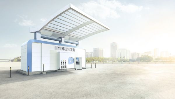 Eine weiße Wasserstofftankstelle steht auf freier Betonfläche mit Bäumen und Hochhäusern im Hintergrund.