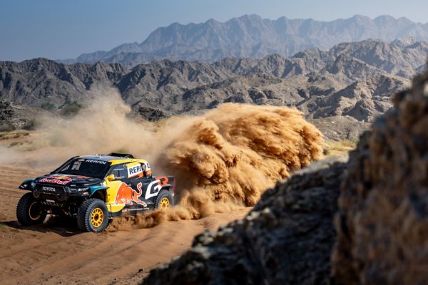 Ein Rallyeauto wirbelt eine Staubwolke auf, während es über eine unbefestigte Strecke in einer bergigen Wüstenlandschaft rast.