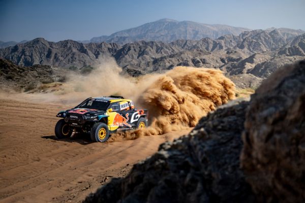 Een Toyota GR DKR Hilux Evo racet door het zand in een spectaculair berglandschap.