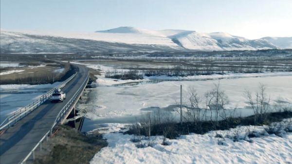 Vogelperspektive eines silbernen Autos mit Dachbox, der über eine Brücke fährt durch eine Winterlandschaft mit Gebirgen im Hintergrund.