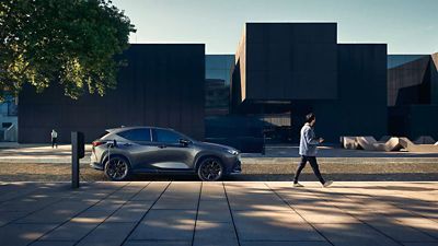 Seitenansicht eines grauen Lexus, der an einer Ladestation vor einem modernen, dunklen Gebäude geparkt ist; im Hintergrund läuft eine Person über den Platz.