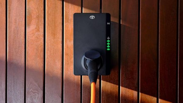 Eine fest installierte Wallbox von Toyota ist an einer Holzfassade montiert und zeigt grüne LED-Anzeigen während des aktiven Ladevorgangs. Die Szene veranschaulicht das effiziente und sichere Laden eines Elektroautos zu Hause – eine zentrale Option für das E-Auto laden in Österreich.