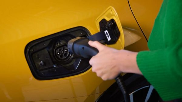 Eine Person in grünem Pullover steckt ein schwarzes Ladekabel in den Ladeanschluss eines gelben Elektroautos. Die Nahaufnahme zeigt den Ladevorgang an der Fahrzeugseite, kurz vor dem Einrasten des Typ2-Steckers. Das Bild steht symbolisch für das einfache und alltagstaugliche Laden von E-Autos.