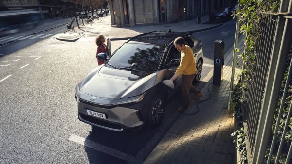 Ein Mann lädt ein Elektroauto an einer öffentlichen Ladesäule in einer sonnigen Stadtstraße, während eine Frau die Beifahrertür öffnet.