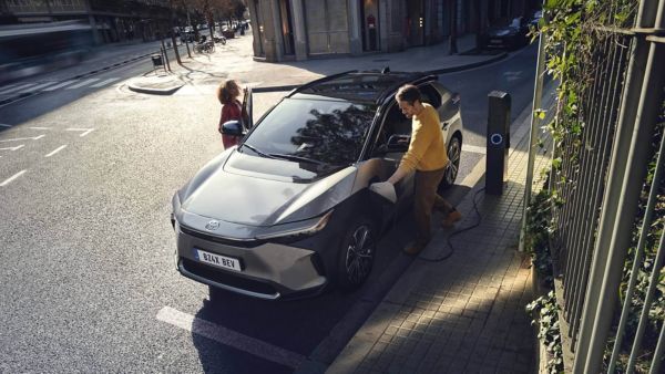 Ein Mann lädt ein silbernes Elektroauto vom Typ Toyota bZ4X an einer öffentlichen Ladesäule am Straßenrand, während eine Frau auf der Beifahrerseite einsteigt. Die Szene spielt bei Tageslicht in einer ruhigen Innenstadtstraße mit moderner Architektur und viel natürlichem Licht. Das Bild veranschaulicht das alltagstaugliche Laden eines E-Autos im öffentlichen Raum an einer öffentlichen Ladesäule.