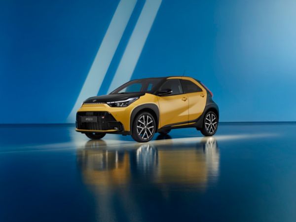 Toyota Aygo X Hybrid colore oro metallizzato con cofano e tetto nero bi-tone, in uno studio blu vivace.