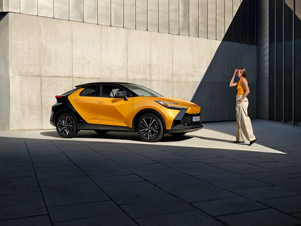 Eine Frau steht vor einem gelb-schwarzen Toyota C-HR, der neben einer modernen Betonfassade geparkt ist.