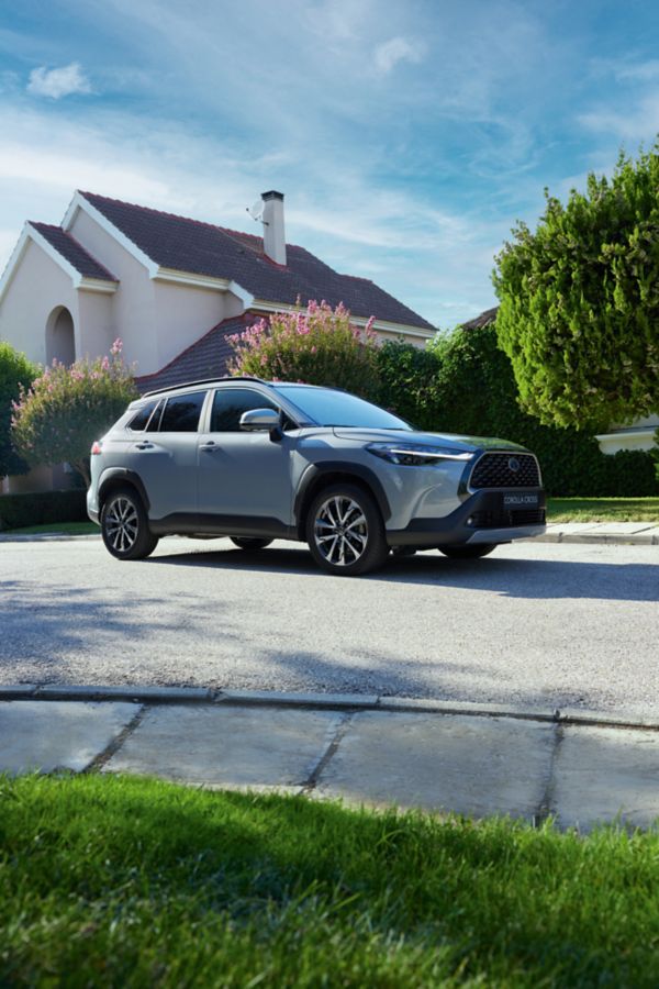 Ein grauer Toyota Corolla Cross steht auf einer ruhigen Straße vor einem Wohnhaus mit Garten und blühenden Sträuchern.