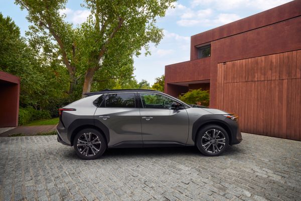 Ein silbergrauer Toyota bZ4X ist vor einem modernen Haus mit roten Fassaden und großen Bäumen geparkt.