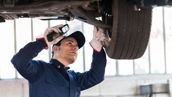 Ein Mechaniker prüft bei einer Auto-Inspektion mit einer Taschenlampe das Fahrwerk eines angehobenen Autos.
