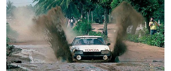 Foto von 1979: Ein weißes Rallye-Auto fährt auf einer schlammigen Straße durch eine Pfütze, sodass das Wasser an den Seiten hochspritzt
