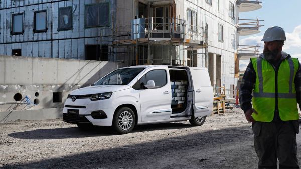 Weißer Proace City mit geöffneter Seitentür auf einer Baustelle, darin die Ladung, im Vordergrund ein Bauarbeiter mit Helm und Warnweste