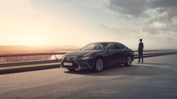 Ein eleganter schwarzer Lexus, der bei Sonnenuntergang auf einer Panoramastraße geparkt ist, und eine Person, die in der Nähe steht und in die Ferne blickt.