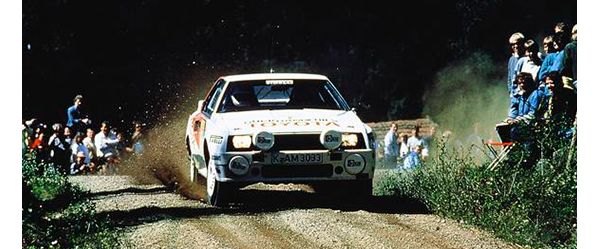 Foto von 1975: Ein weißes Rallye-Auto fährt auf einer Schotterpiste über eine Kuppe und wirbelt Staub auf, neben der Strecke einige Zuschauer