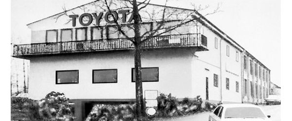 Schwarz-Weiß-Foto eines zweistöckigen Industriegebäudes mit Toyota Schriftzug am Giebel, davor parkende Autos
