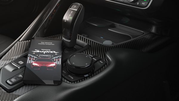 Ein Smartphone liegt auf der Mittelkonsole eines Fahrzeugs, auf dem Bildschirm kann man einen Sportwagen und den Text Toyota Supra Connect sehen