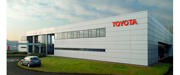 Flache graue Industriehalle mit rotem Toyota Schriftzug, vor dem verglasten Eingangsbereich ein geparktes Auto