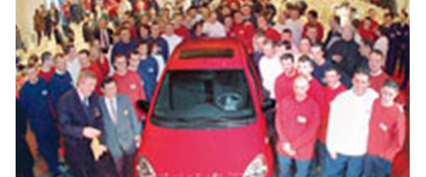Etwa 50 Personen um einen roten Toyota Yaris herum, von etwas erhöhter Position in einer Autofabrik fotografiert