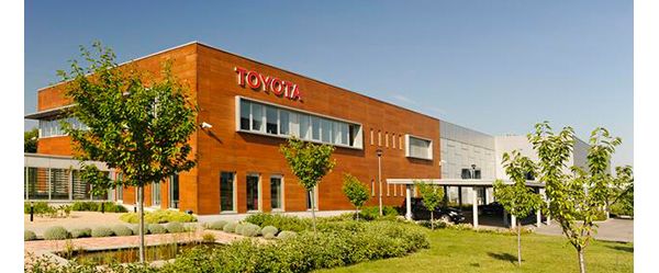 Modernes zweistöckiges Bürogebäude, ockerfarben mit rotem Toyota Schriftzug, davor Büsche und Bäume