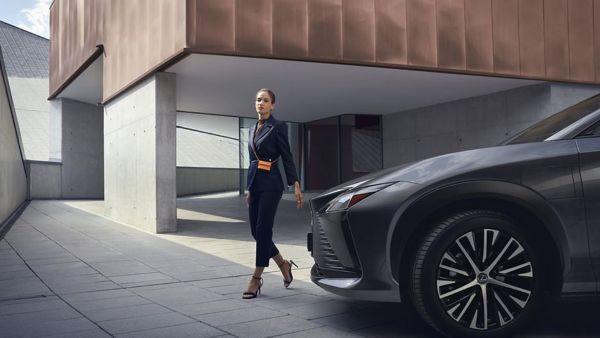 Eine Person in eleganter Kleidung geht selbstbewusst auf einen schwarzen Lexus zu, der in einer modernen städtischen Umgebung geparkt ist. 