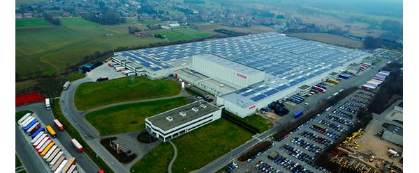 Luftaufnahme einer großen Autofabrik mit rotem Toyota Schriftzug, daneben ein Parkplatz mit Autos und LKWs, im Hintergrund eine kleine Stadt