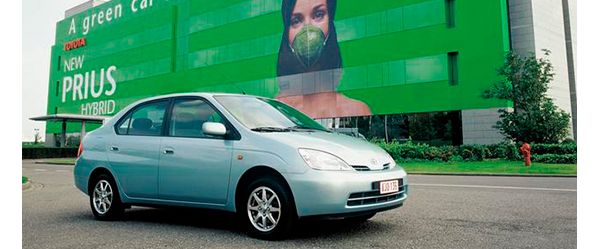 Blaugrauer Toyota Prius von 2000 vor einem Toyota Gebäude mit grün bedruckter Fassade, darauf eine Frau mit grünem Mund-Nasen-Schutz und die Aufschrift „A green car, New Prius Hybrid“