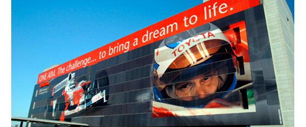Überdimensionales Poster an einer Hauswand, darauf ein Formel-1-Rennwagen und ein Fahrer mit Helm im Cockpit, Schriftzug: „The challenge … to bring a dream to life.“