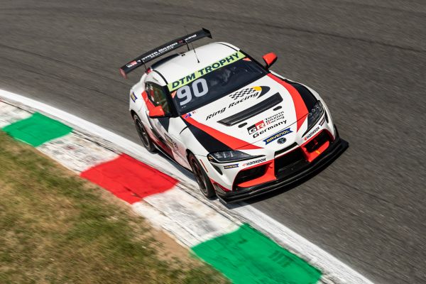 Weißer GR Supra GT4 mit schwarz-roter Folierung und Sponsoren-Logos fährt bei einem Rennen durch eine Kurve, leicht erhöht von vorn fotografiert