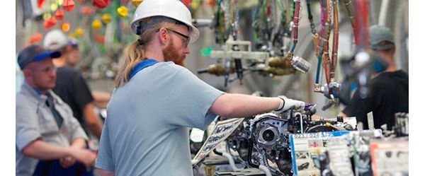 Ein Fabrikarbeiter mit Helm, Gehörschutz und Handschuhen arbeitet an einem Fahrzeugbauteil, im Hintergrund diverse Werkzeuge und weitere Arbeiter