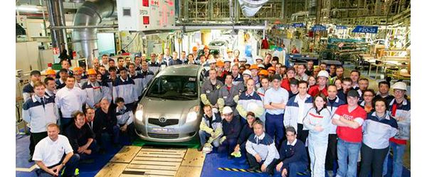 Etwa 50 Personen um einen silbernen Toyota Yaris herum, von etwas erhöhter Position in einer Autofabrik fotografiert
