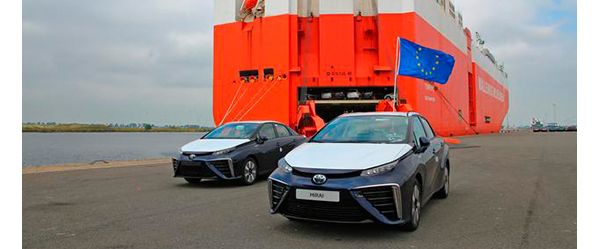 Zwei blaue Toyota Mirai von 2015 vor einem großen Containerschiff, an einem der Autos ist eine Europaflagge befestigt