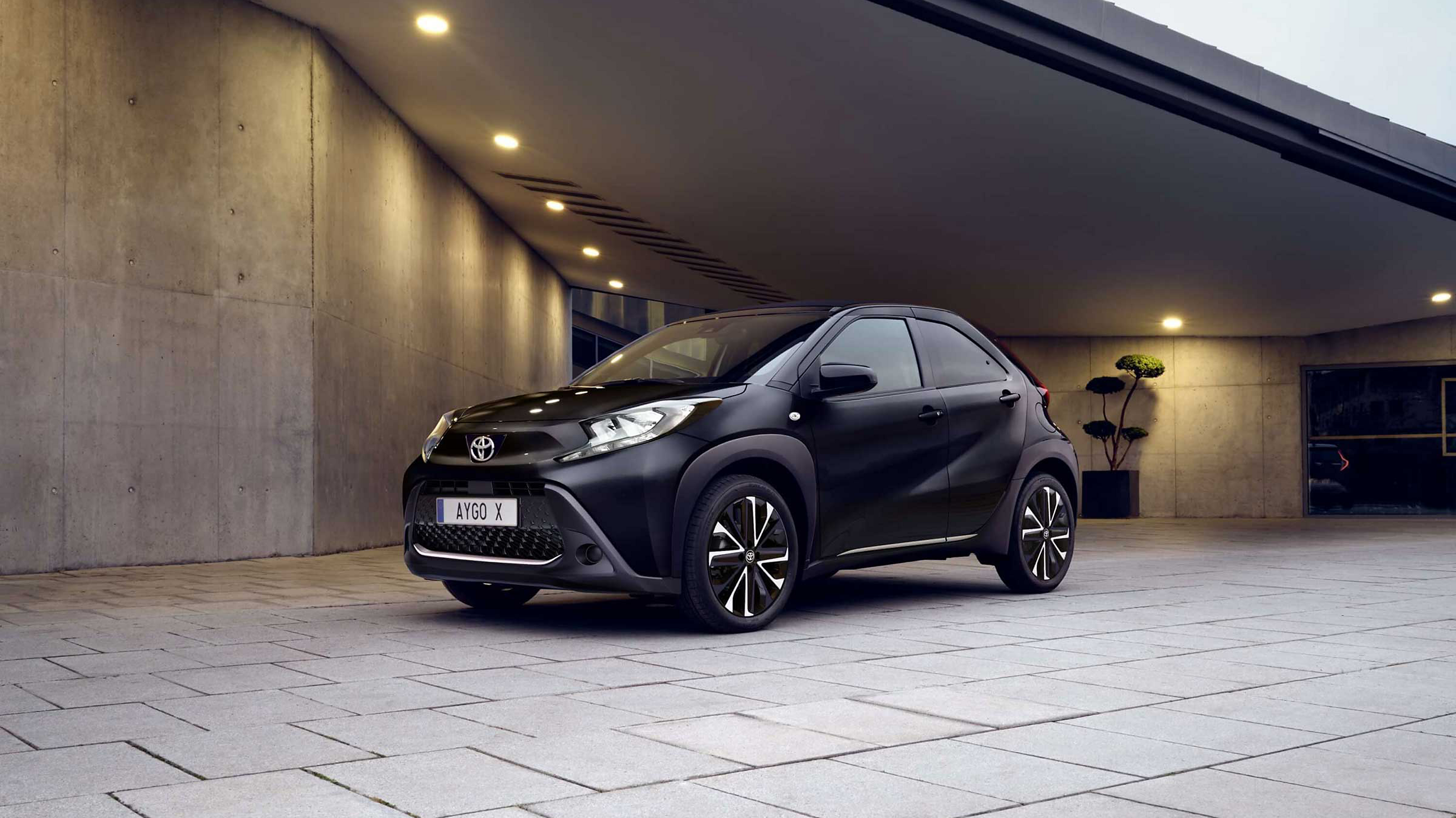 Seitliche Frontansicht eines schwarzen Aygo X, der unter einer beleuchteten Überdachung eines modernen Gebäudes steht.