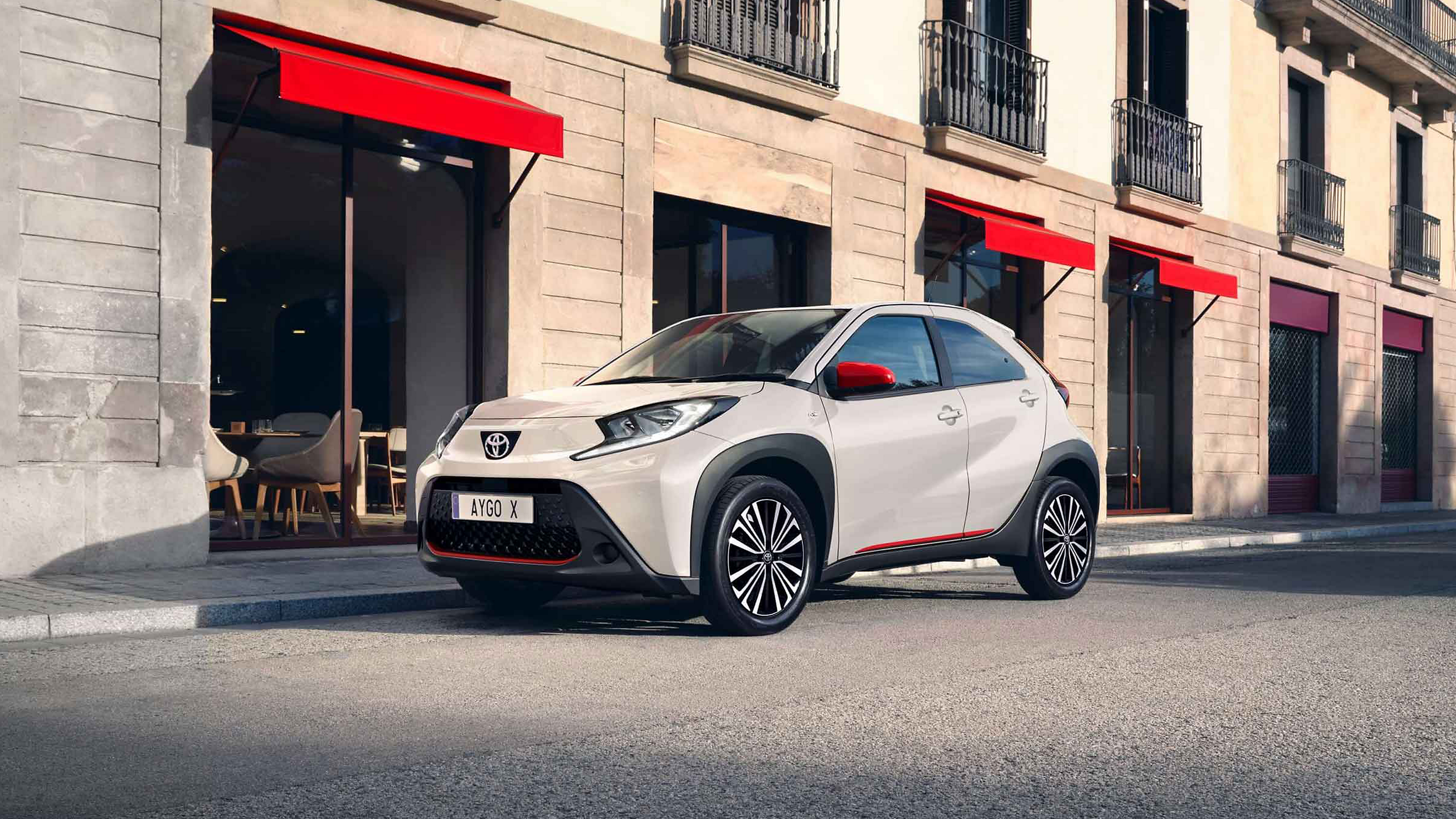 Seitliche Frontansicht eines weißen Aygo X mit roten Akzenten, der an einer Stadtstraße geparkt ist mit roten Markisen über Schaufenster im Hintergrund.